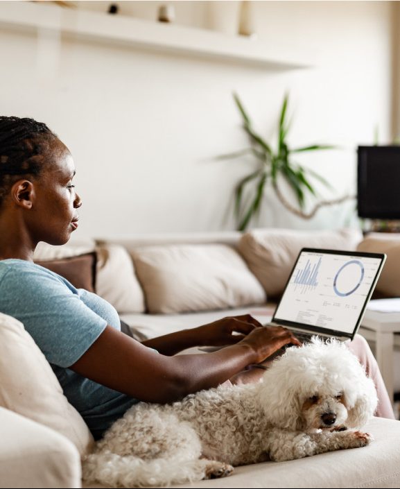 A Black woman sits on her couch, with a dog beside her and a laptop on her lap, indicating she might be pursuing one of many ways to make money at home.