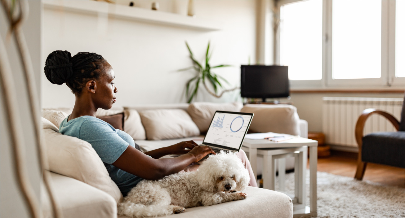 A Black woman sits on her couch, with a dog beside her and a laptop on her lap, indicating she might be pursuing one of many ways to make money at home.