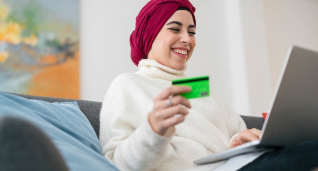 From below of joyful Muslim female in hijab sitting on couch with laptop and plastic card while making purchase during online shopping