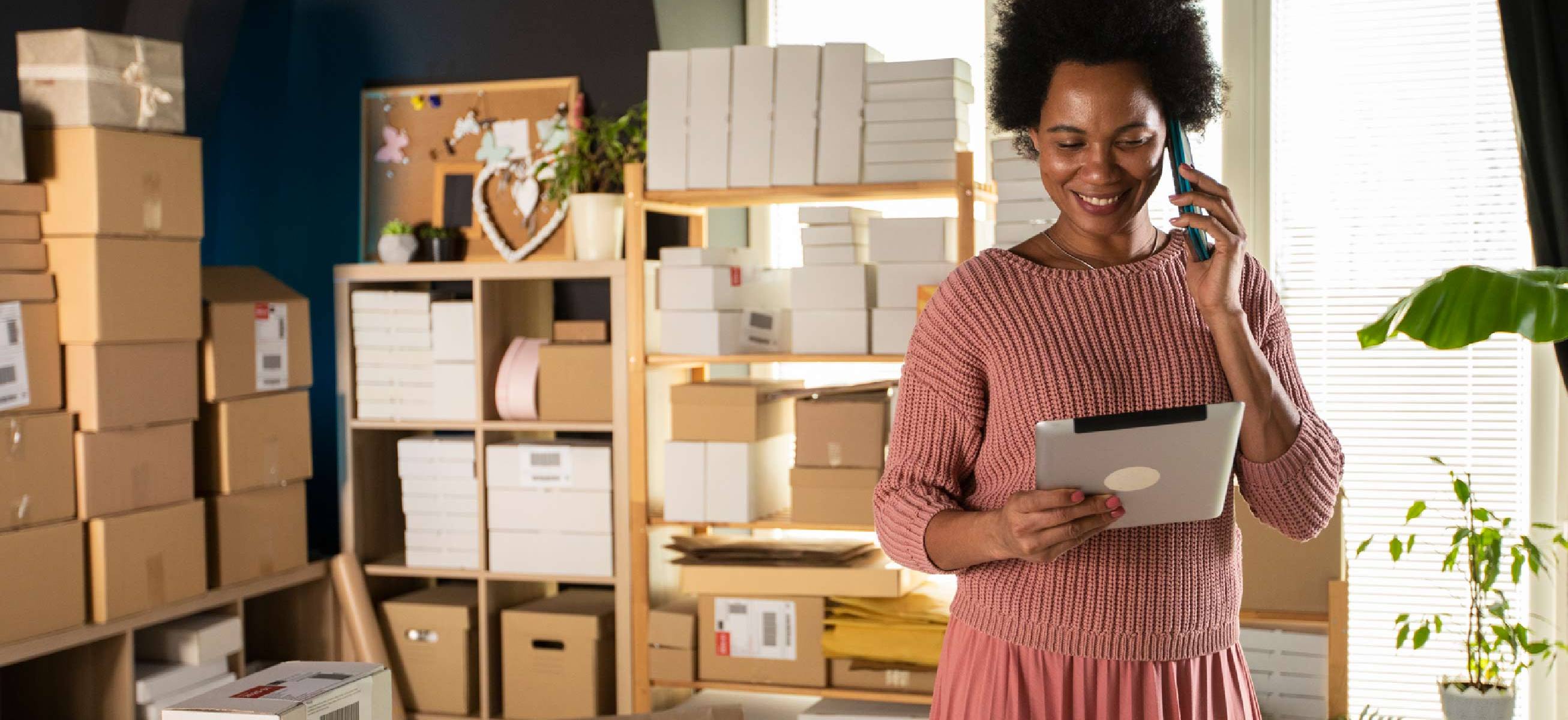A Black woman and small business owner is in a shop surrounded by packages, looking at her tablet, indicating she might be using the cost of goods sold formula for her accounting.