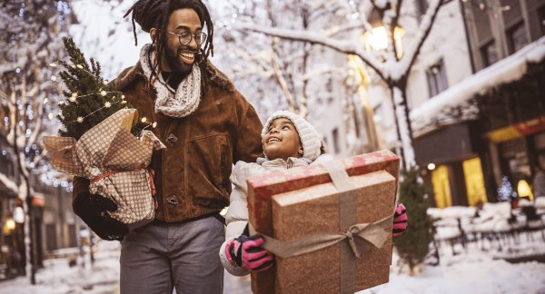 A Black man holds a small Christmas tree walking in a snowy city alongside a kid holding two gift boxes while smiling at each other, indicating they have their holiday budgeting in order.