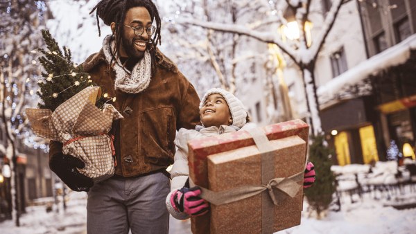 A Black man holds a small Christmas tree walking in a snowy city alongside a kid holding two gift boxes while smiling at each other, indicating they have their holiday budgeting in order.