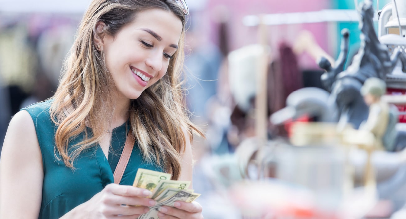 A woman is counting the cash she has in her hand while smiling at objects for sale, indicating she’s using her fiat money to buy something.