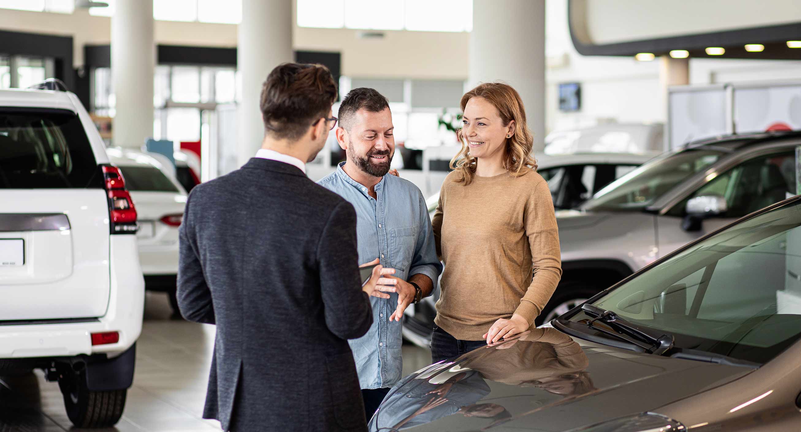 A couple talks to a salesman at a car dealership, indicating they are buying a car that’s within their budget in order to start saving money.