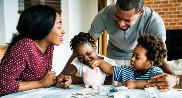 A Black family gathers around a table, with a piggy bank and cash sitting in front of them, indicating they might be attempting a money-saving challenge.