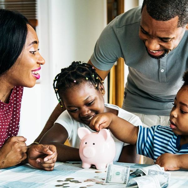 A Black family gathers around a table, with a piggy bank and cash sitting in front of them, indicating they might be attempting a money-saving challenge.