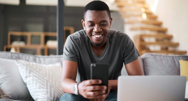 A Black man sits on his couch in front of his computer while looking at his phone and smiling indicating he is saving money.