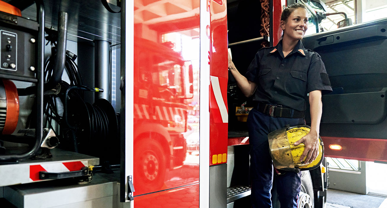 A firefighter stands on the steps of the fire truck and smiles as she looks ahead, suggesting that she considers her job one of the most fulfilling jobs.