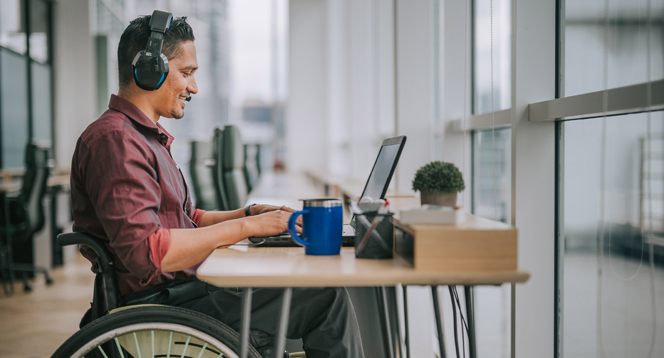 A man uses a wheelchair while sitting at his work desk during an online meeting. He smiles at the screen indicating he’s happy to have one of the most fulfilling jobs.
