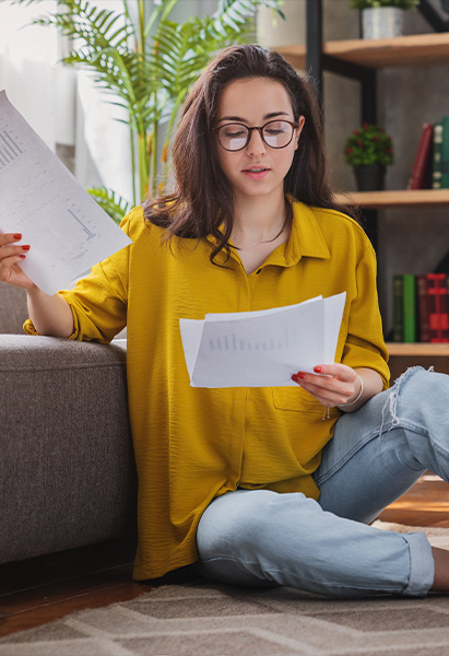 A woman in a yellow blouse sits on the floor of her living room reviewing documents, indicating she might be researching student loan forbearance.