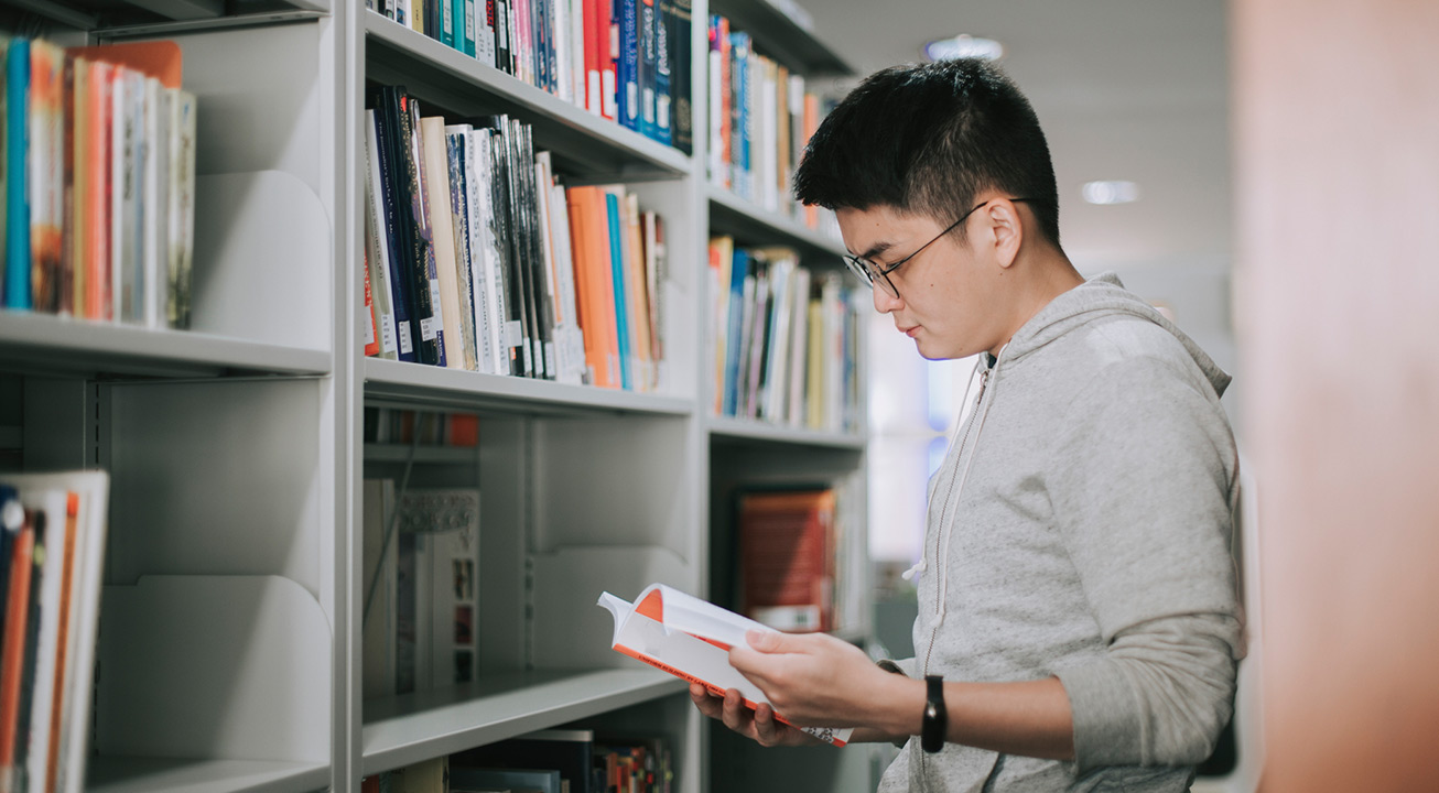 Student reading a book in a library in front of the shelves full of books.